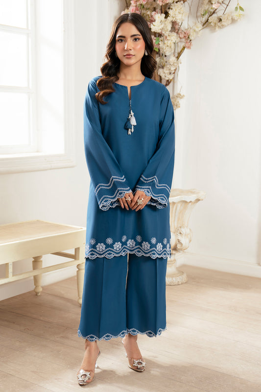 Woman wearing a blue dress with white embroidery in a room with a white wall and wooden floor.