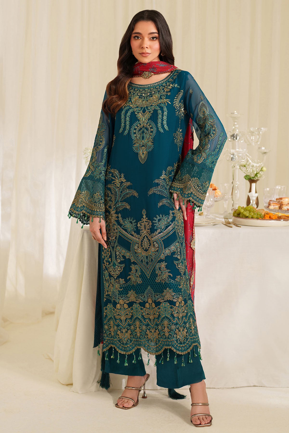 Woman wearing a teal embroidered traditional outfit with a red scarf in a decorated room.