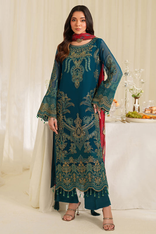 Woman wearing a teal embroidered traditional outfit with a red scarf in a decorated room.