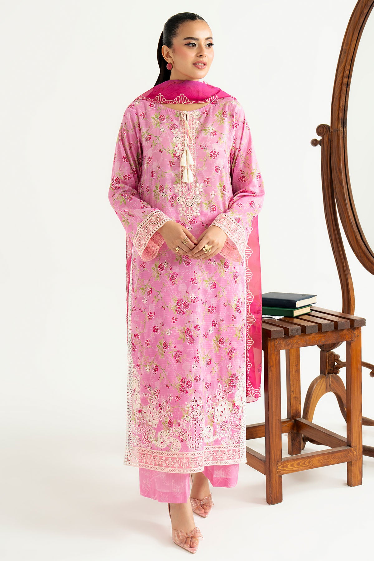 Woman wearing a pink traditional outfit with floral patterns, standing next to a wooden stool and mirror.
