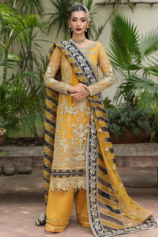 Woman in a traditional yellow and black embroidered outfit with a matching dupatta, standing outdoors with plants in the background.