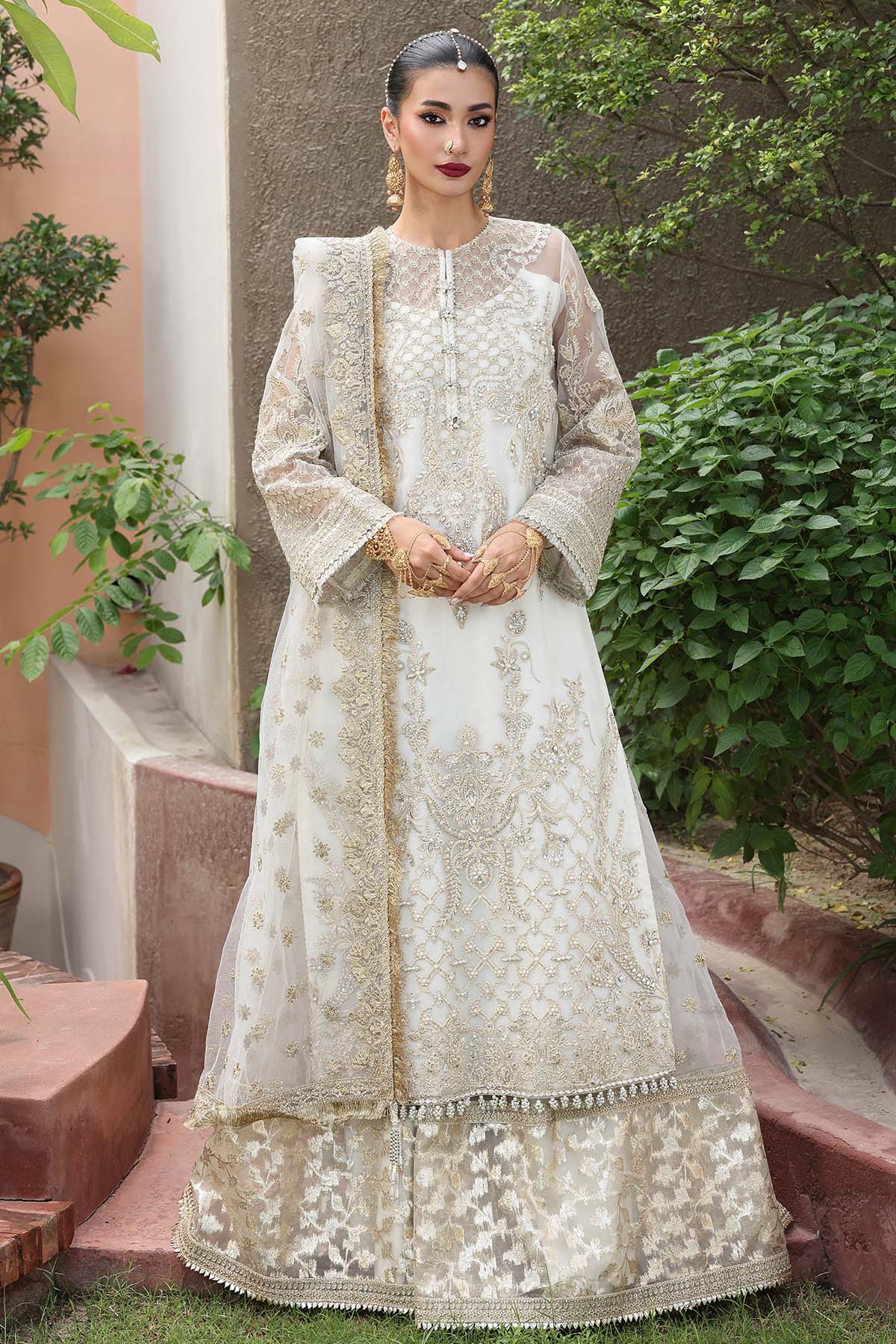 Woman in a white embroidered traditional outfit standing outdoors with greenery.