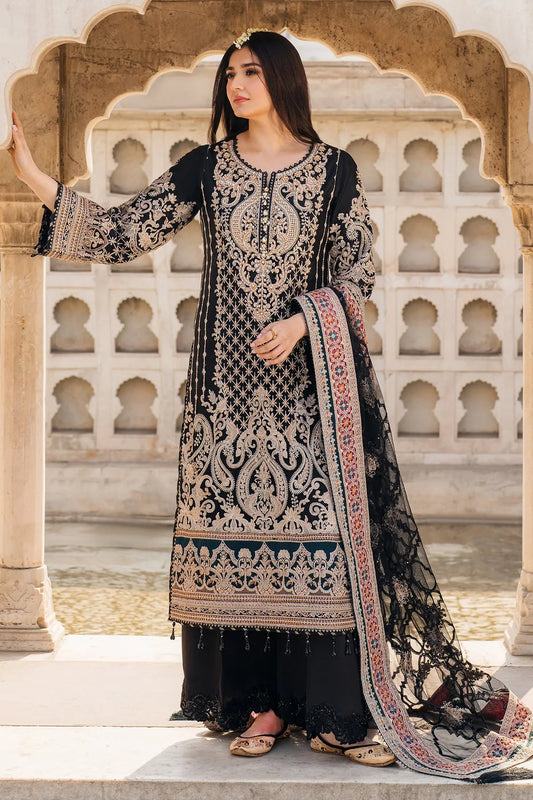 A woman wearing a black, red, and green embroidered three-piece clothing ensemble, consisting of a net shirt with plain back and sleeves, a net dupatta, and raw silk trousers, standing against an archway background.