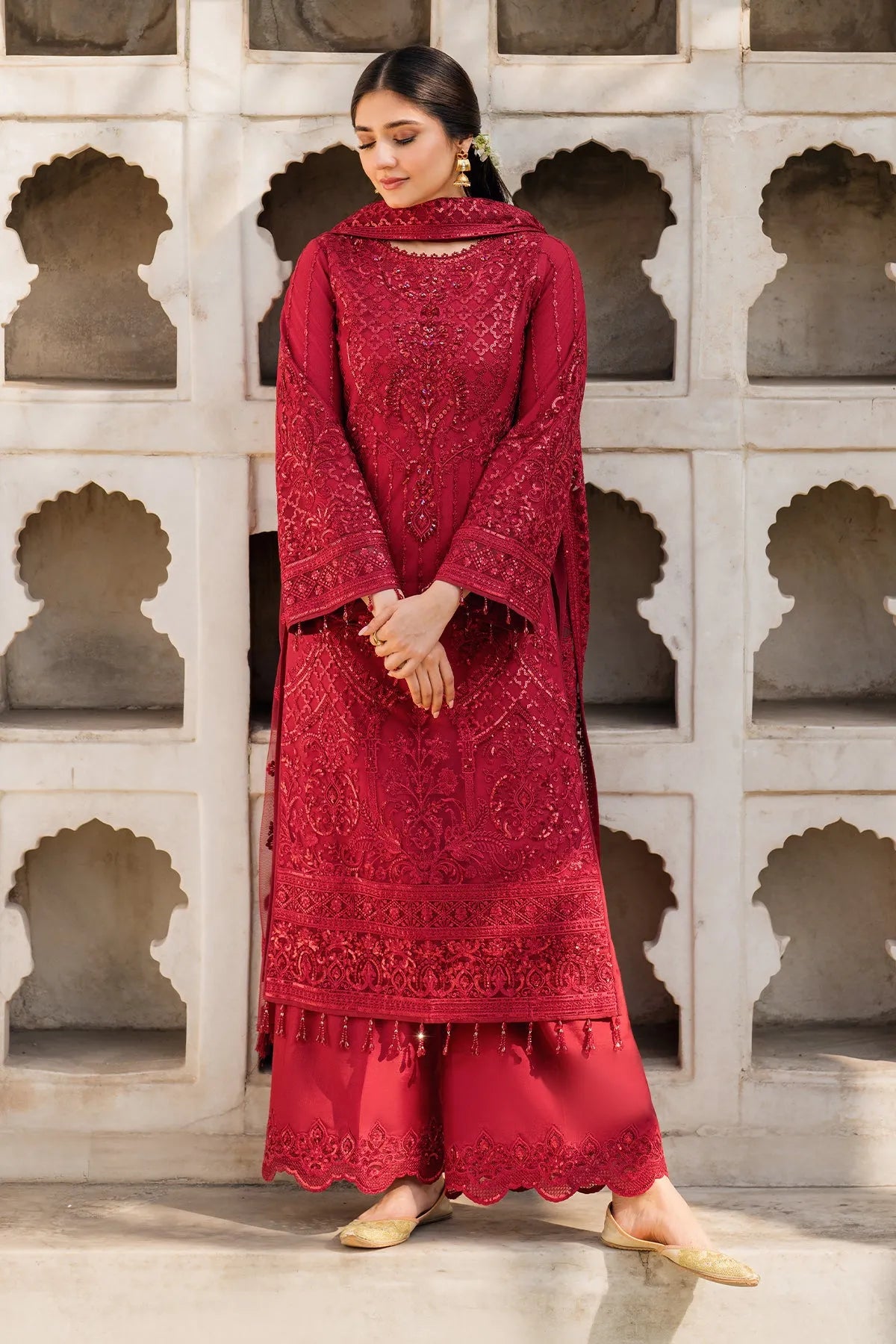 A woman wearing a red, three-piece ensemble with intricate embroidery, featuring a net front, plain net back, and sleeves with embroidered organza details. Accompanied by a net dupatta and a dyed raw silk trouser with embroidered organza border.