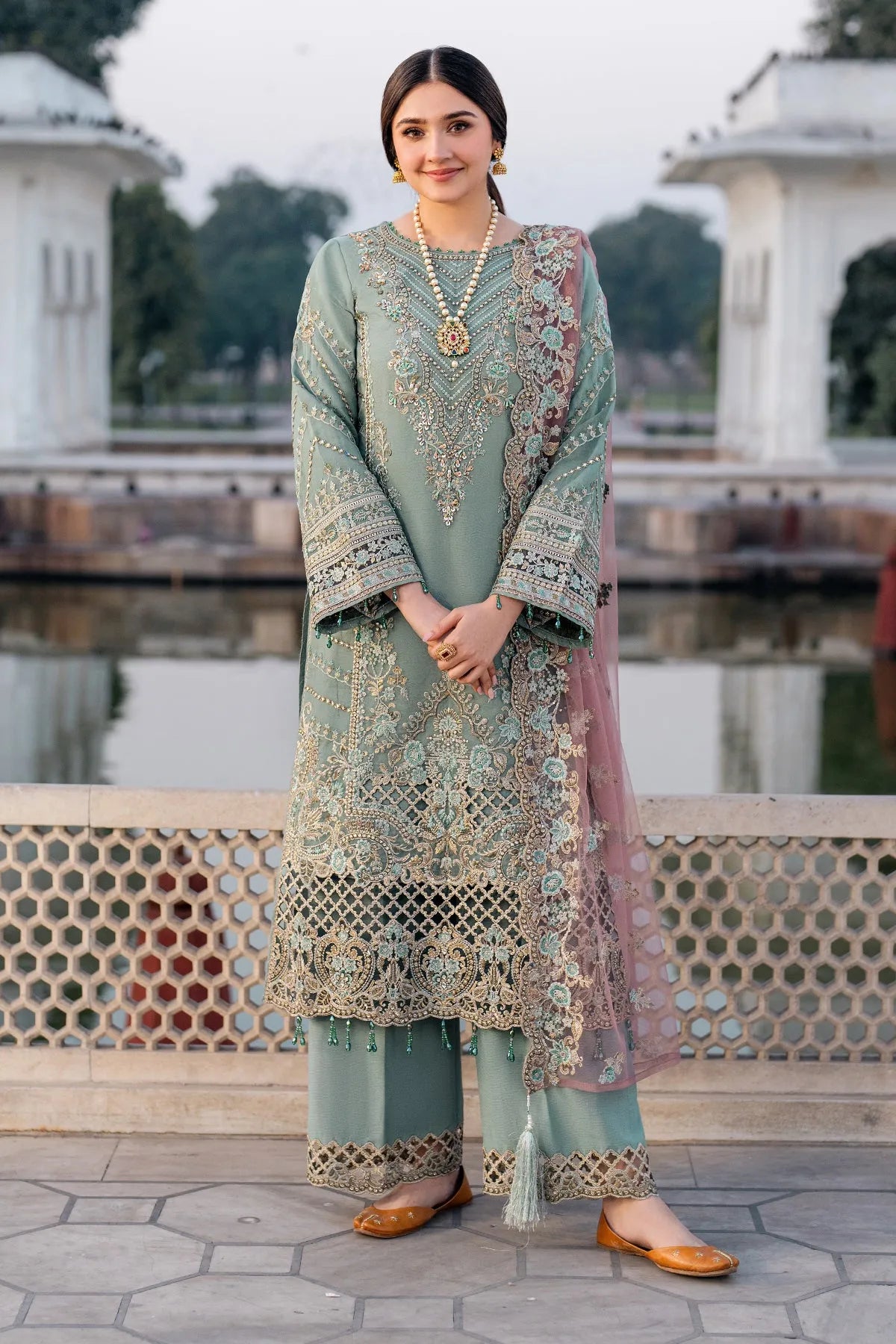 A woman wearing a green and pink traditional outfit with embroidery and an organza dupatta, standing near a water body.