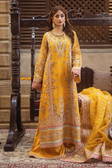 A woman wearing a traditional yellow embroidered gown with handmade embellishments, paired with a dupatta and sitting next to a couch with a decorative drape.
