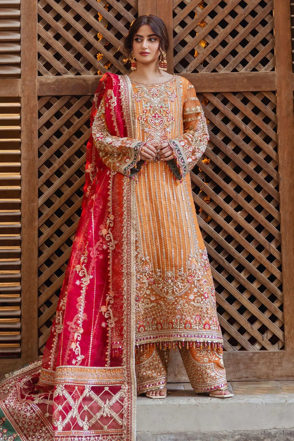 A woman wearing a traditional, embroidered red and orange outfit with a dupatta, standing in front of a paisley patterned backdrop.