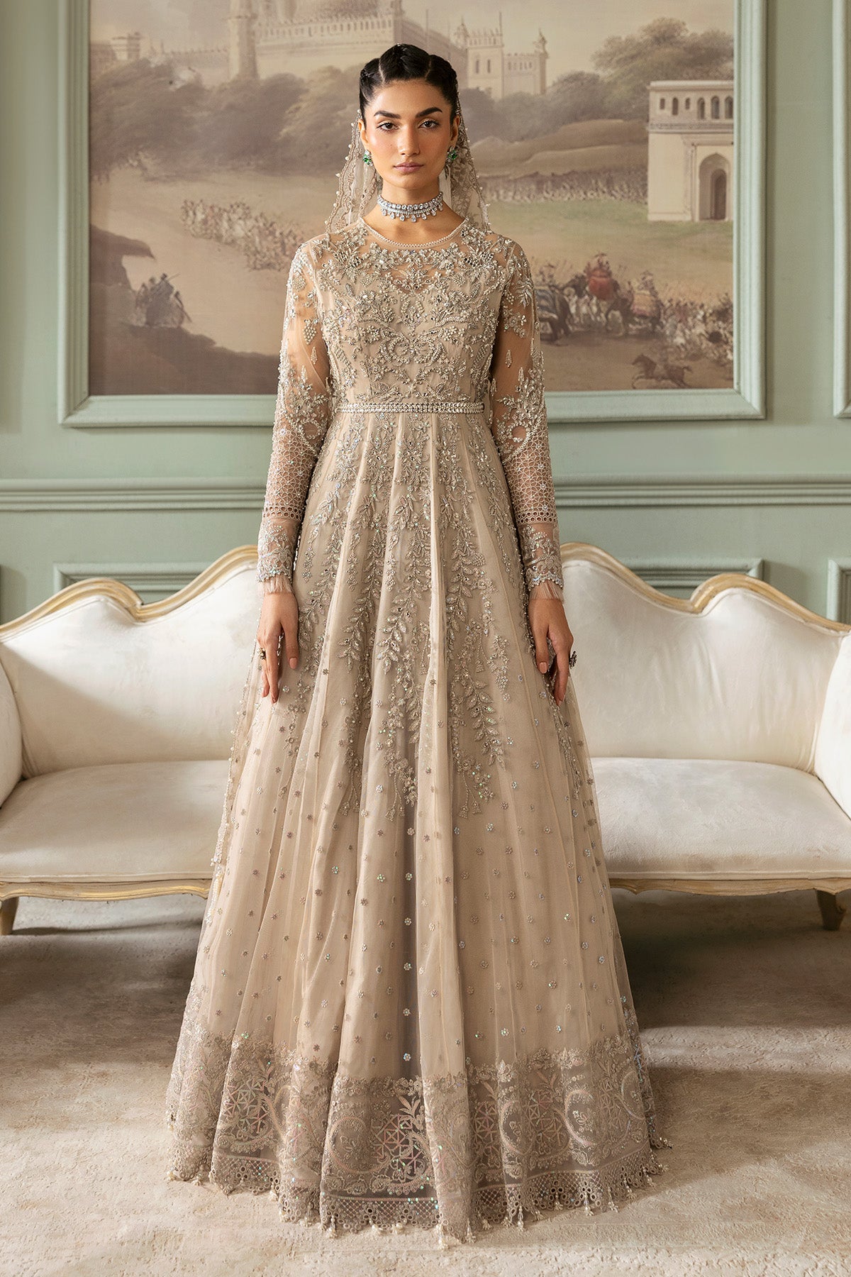A woman wearing a beige and silver embroidered gown with long sleeves and a floor-length skirt, accessorized with a net dupatta.