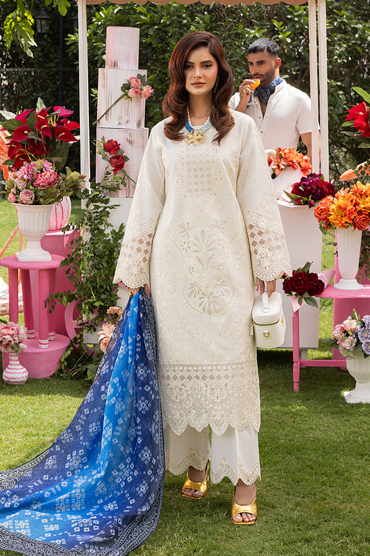 A woman wearing a white traditional Pakistani dress with intricate embroidery, accompanied by a blue printed silk dupatta and white trousers, posing outdoors with floral decorations in the background.