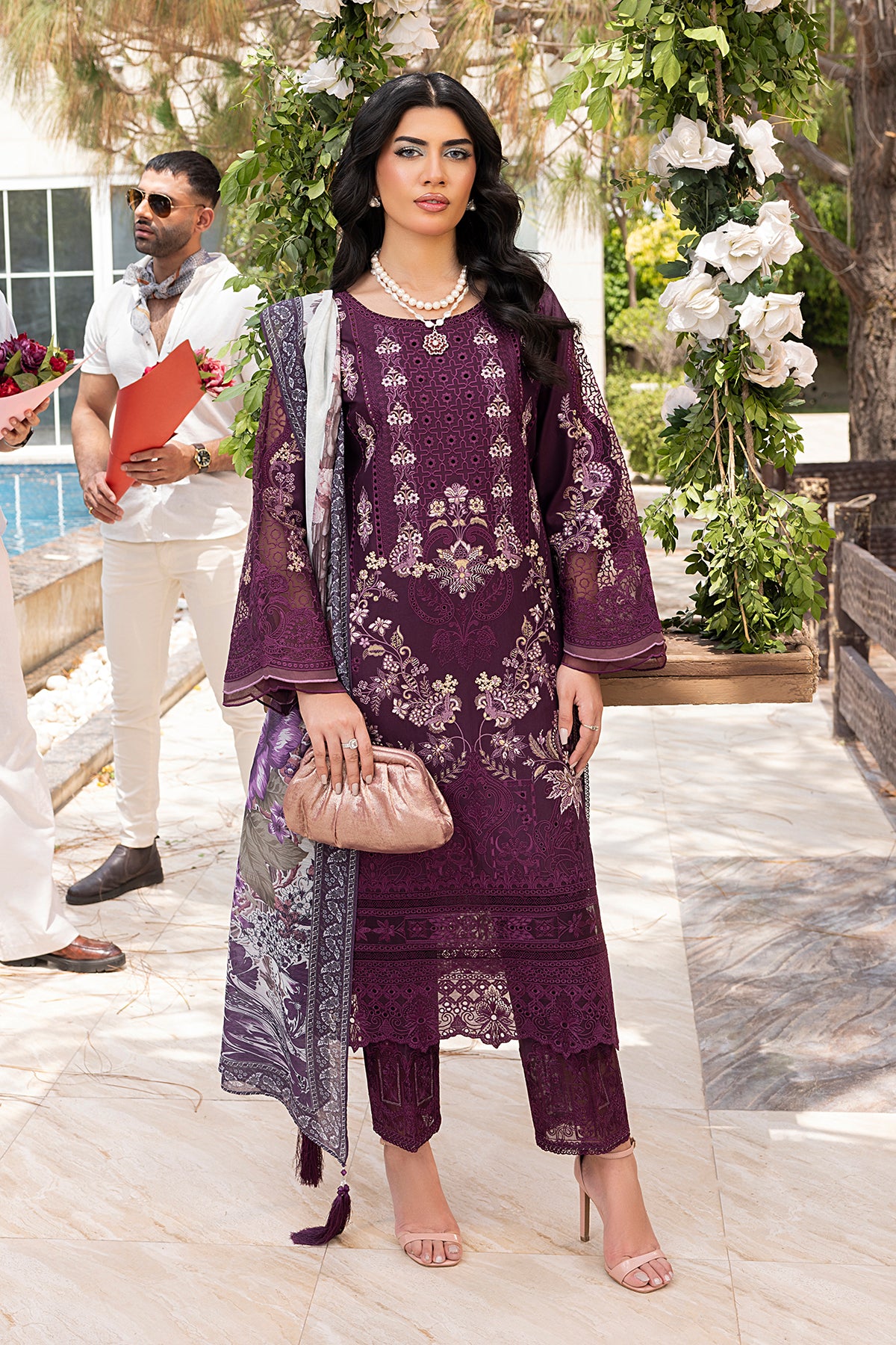 A woman wearing a traditional Plum-colored outfit with detailed embroidery, consisting of a shirt, dupatta, and trousers, standing in a garden setting.