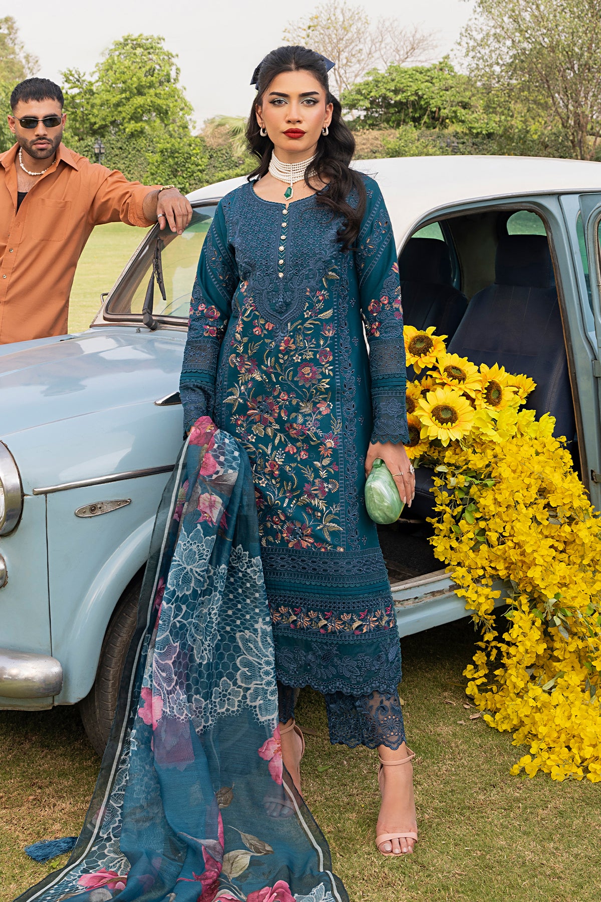A woman standing beside a vintage car, wearing a teal blue traditional outfit with floral embroidery, including a shirt, dupatta, and trousers.