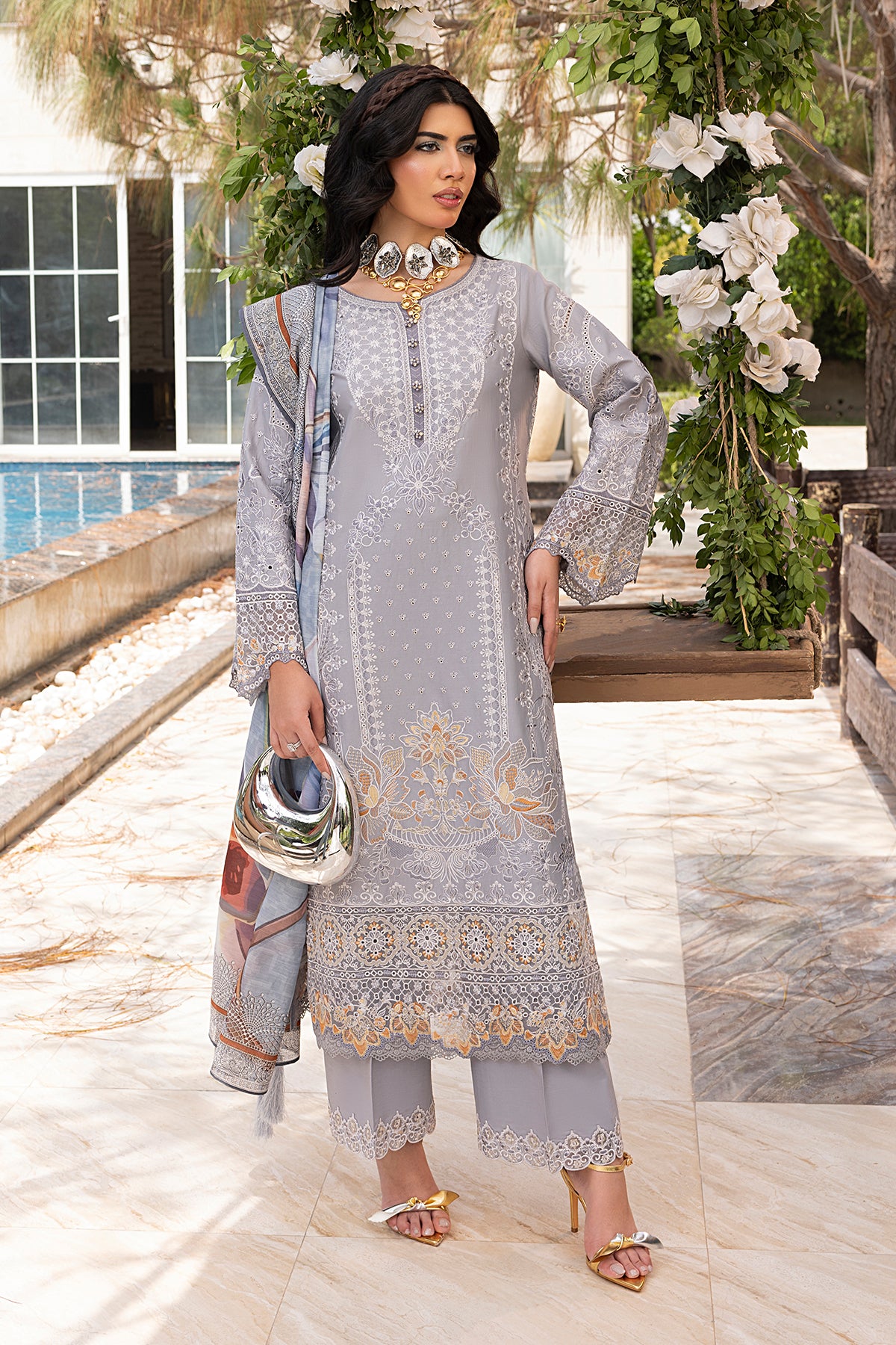 A woman wearing a traditional grey outfit with intricate embroidery, light trousers, and a dupatta with a print. She is standing near a pool with a floral backdrop.