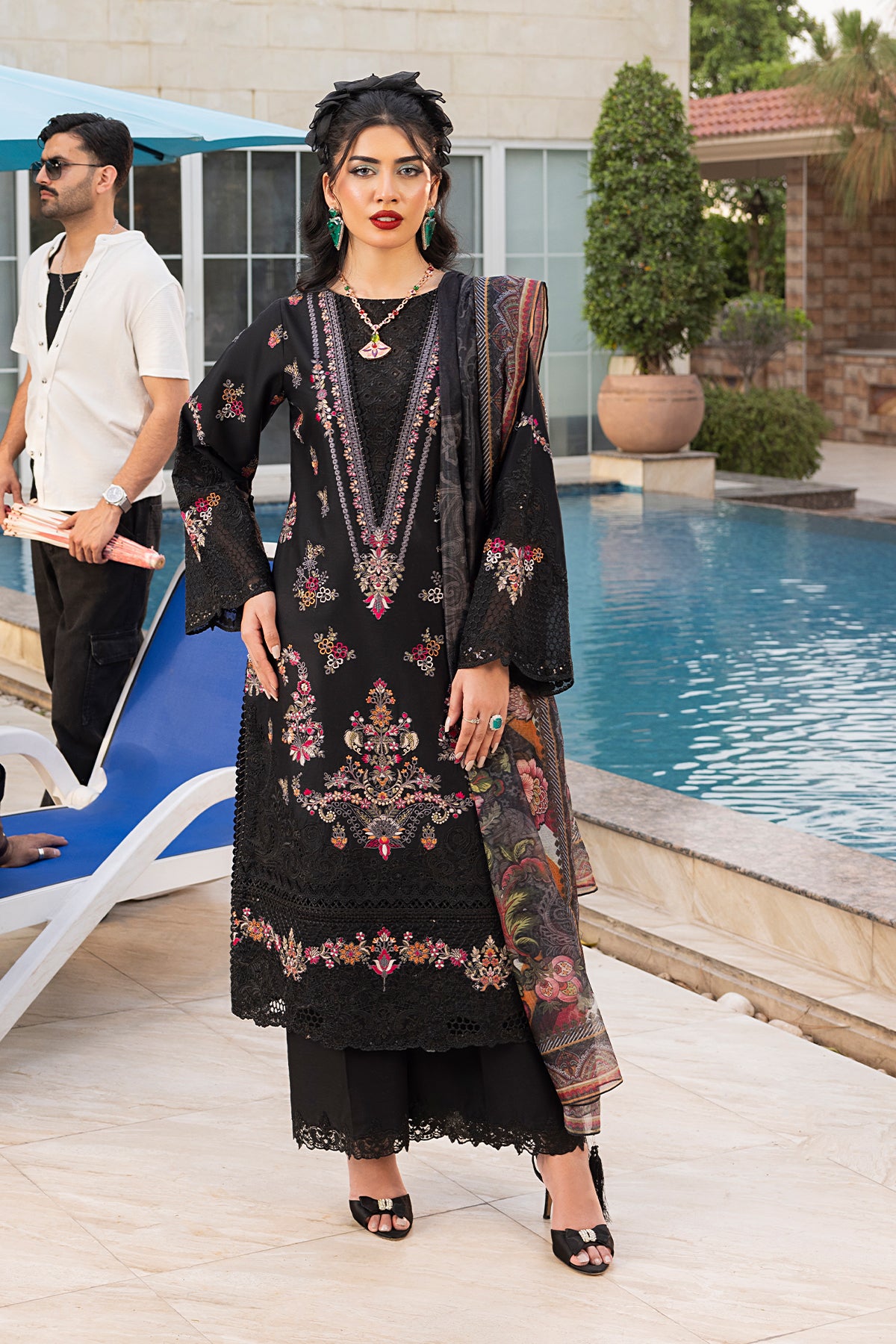 A woman standing by a pool wearing a black traditional outfit with red and white embroidery, including a shirt, dupatta, and trousers.