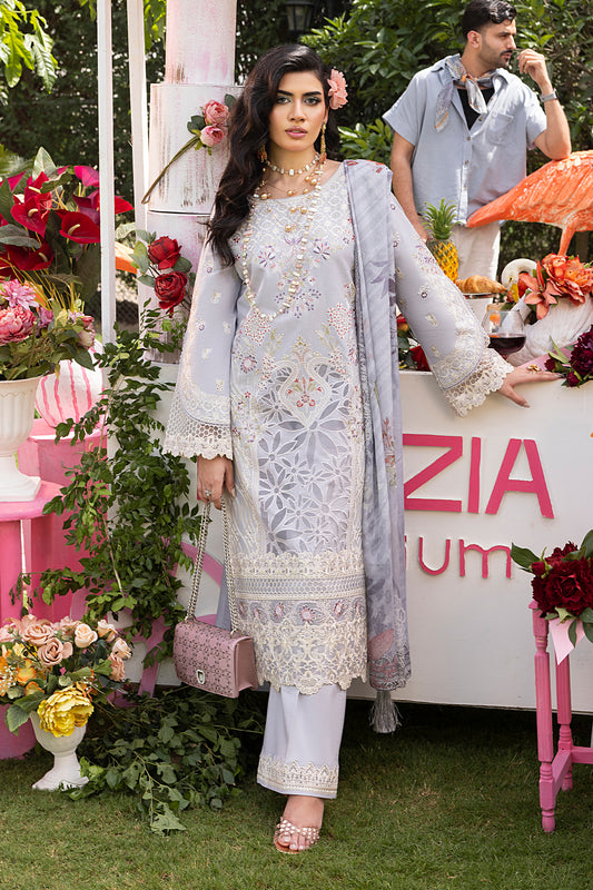 A woman wearing a white and blue embroidered cotton dress with a dupatta, standing in front of a floral backdrop with a sign that reads 'ZIA'. The dress features intricate motifs and the dupatta has a printed design.